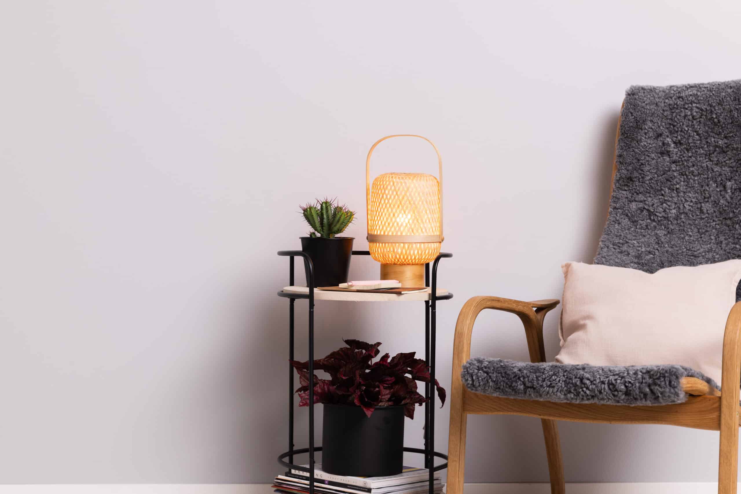 Lampa lyser mjukt på ett bord med växter och tidningar. En fåtölj med grått överdrag och kudde står bredvid, skapar en mysig atmosfär. Förbättra ditt hem med Clas Fixare.