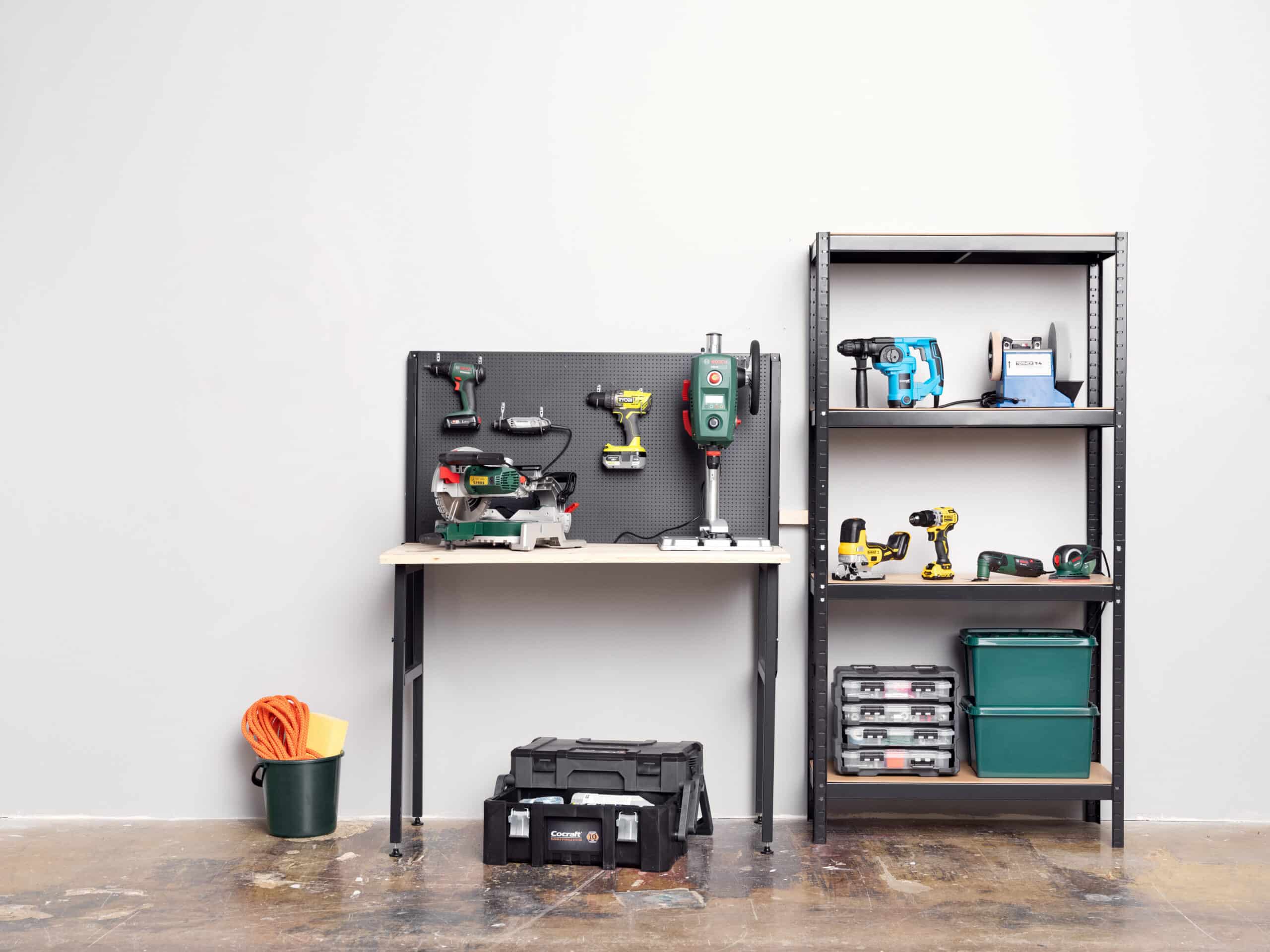 Arbetsbänk med verktyg som sågar och borrar står redo för snickaruppdrag. Hylla bredvid håller fler verktyg för elektrikerjobb. Hink med kabel och svamp ligger på golvet.